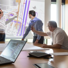 Foto Besprechung Mitarbeiter erklärt Planung Tanklager an Leinwand, mehrere Mitarbeiter diskutieren